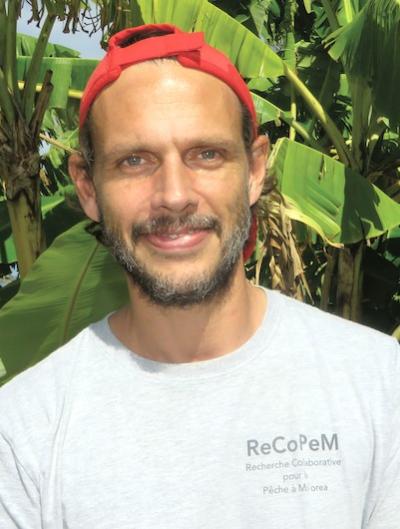 Jean smiles in front of some banana trees with a red backwards hat on